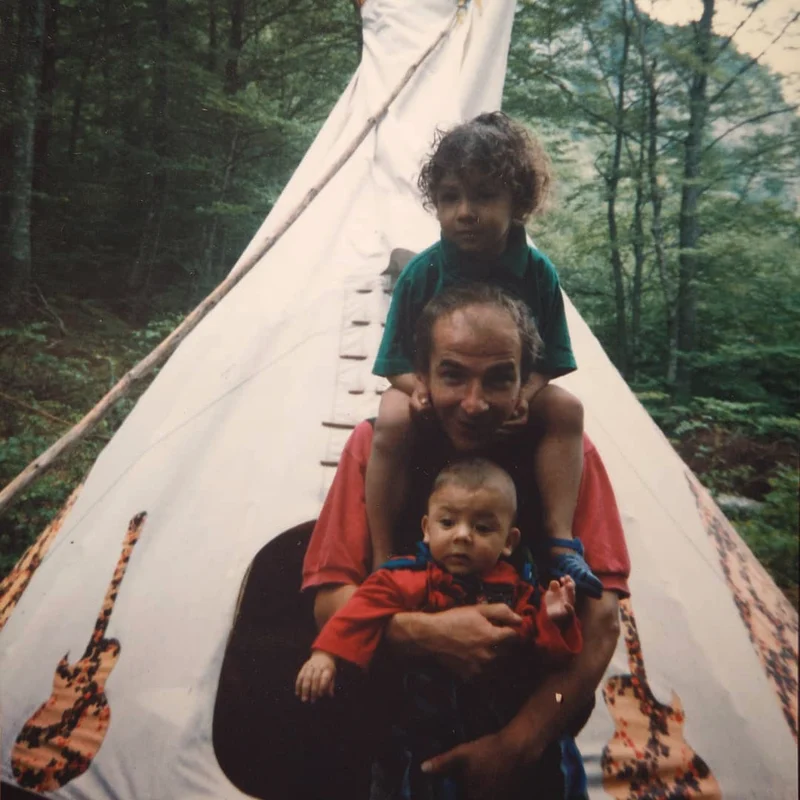 Photo d’archive : le fondateur portant ses deux fils devant un tipi, en forêt